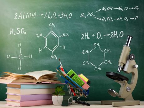 Education and science concept - group of colorful books and microscope on the wooden table in the classroom, blackboard background