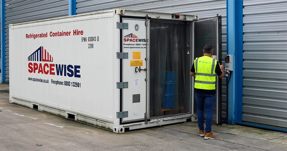A white branded portable refrigeration unit sits outside in a car park, besides rolling shutters. The front door is currently open and a ReAgent worker with a hi-vis jacket is inspecting a list inside the door.
